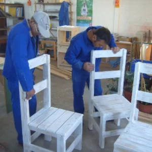 Two men wearing matching blue coveralls are inspecting or working on rough-hewn wooden chairs that have been painted white. They are bending over the chairs in an indoor workshop, which appears to be focused on carpentry or woodworking.