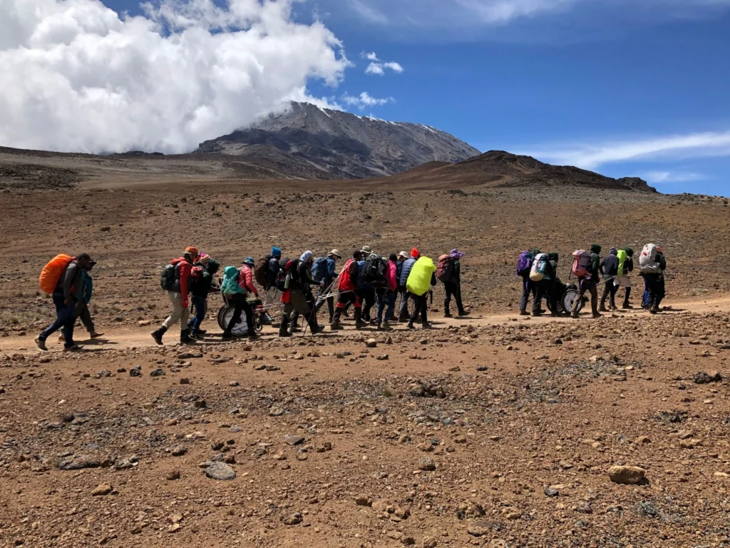 A group of approximately twenty hikers, some of whom are pushing a large, rugged all-terrain wheelchair, trekking across a wide, reddish-brown, desolate plain. They are walking away from the camera toward the snow-capped peak of Mount Kilimanjaro, which is partially obscured by clouds under a bright blue sky.