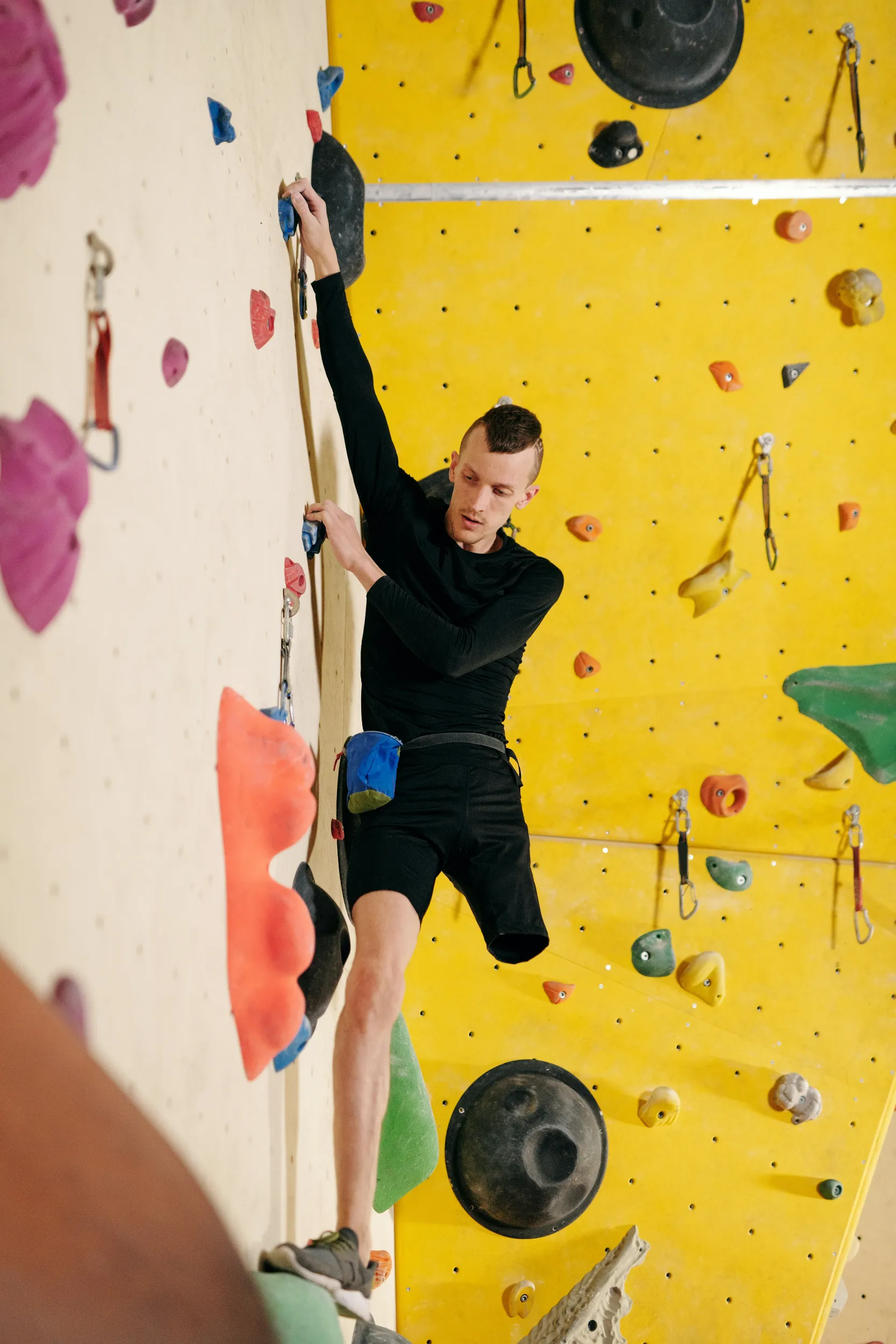 A young man wearing a black long-sleeved shirt and black shorts is climbing an indoor rock wall. He is reaching for a hold with his right hand. He has a lower-limb difference, with his right leg ending above the knee. The wall is light yellow and features various colorful climbing holds and safety equipment.