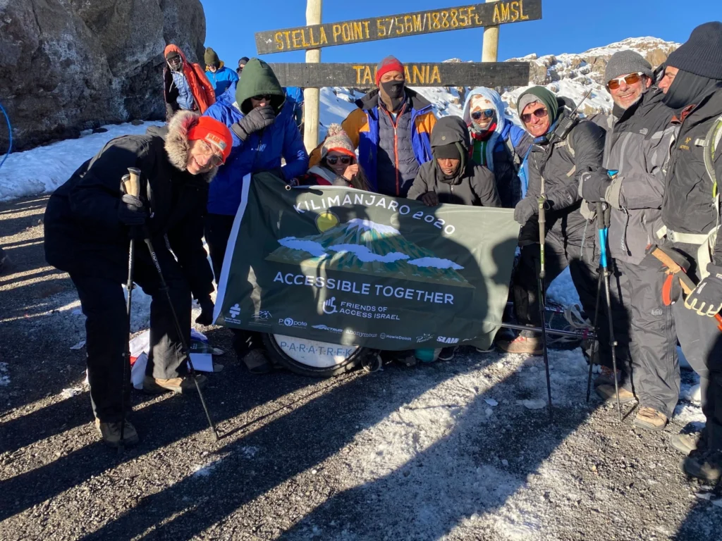A group of hikers bundled in cold-weather gear, holding hiking poles, posing together in the snow at the summit of Mount Kilimanjaro. They are gathered around a wooden sign marking "Stella Point 5756M/18885Ft. AMSL" and holding a banner that reads "KILIMANJARO 2020 ACCESSIBLE TOGETHER" with the logo for Friends of Access Israel.