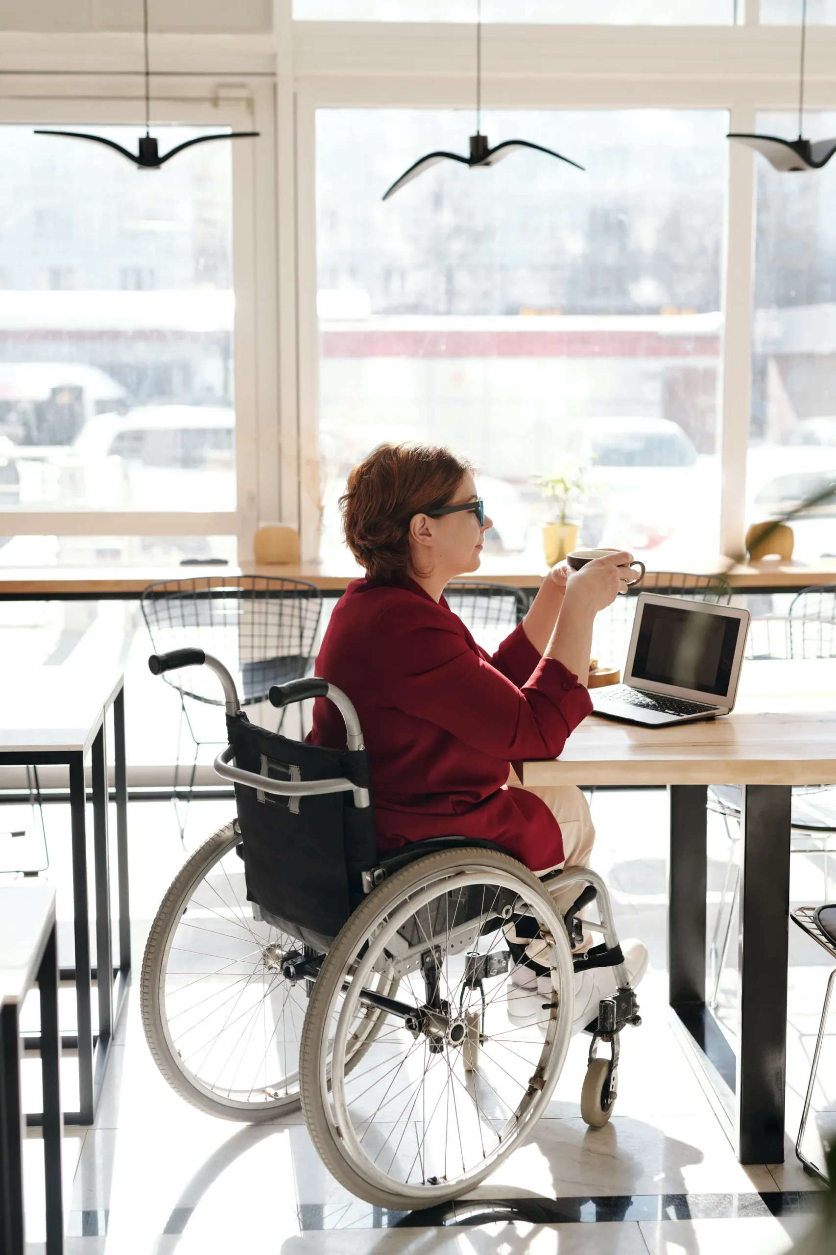 A woman with red hair and glasses, wearing a dark red jacket, is sitting in a wheelchair at a wooden table in a brightly lit cafe or office space. She is looking away from the camera, holding a coffee cup in one hand near an open laptop. Large windows behind her show a bright outdoor scene.