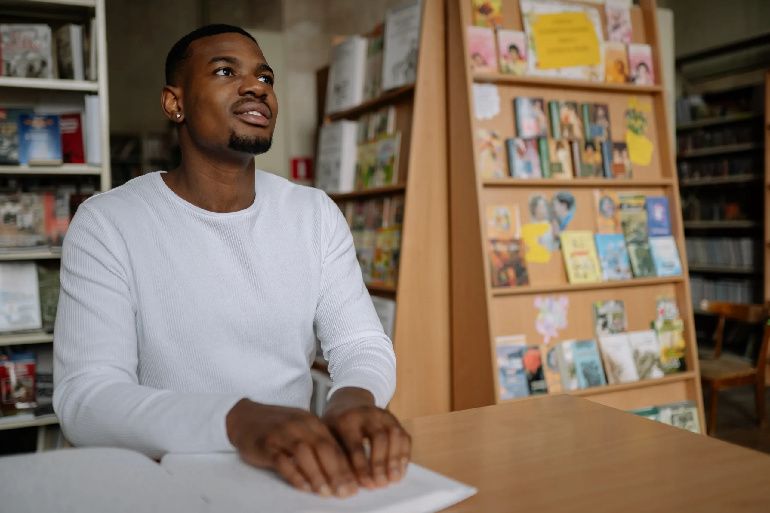 A Black man with a beard, wearing a white long-sleeve shirt, is sitting at a wooden table in a library or bookstore. He is gently touching a book open on the table, indicating he may be reading Braille. He is looking up and slightly away from the camera. Bookshelves are filled with books in the background, including one shelf showcasing small, colorful book covers.