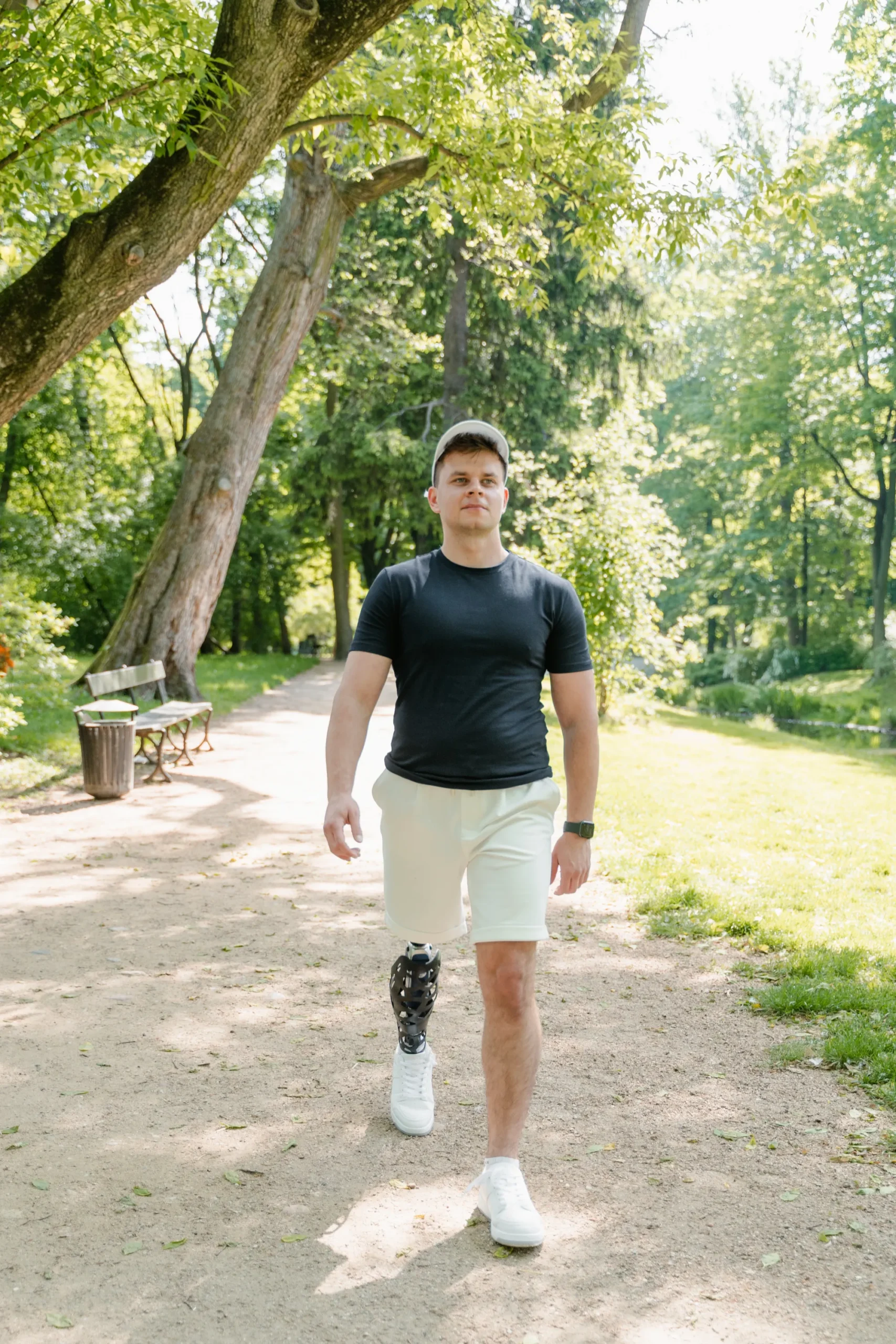 A young man with an above-knee prosthetic leg walks confidently on a dirt path through a sunny park. He is wearing a black t-shirt, light-colored shorts, a baseball cap, and white sneakers. The prosthetic is a distinct black and white mechanical device. Large green trees and a park bench are visible in the background.