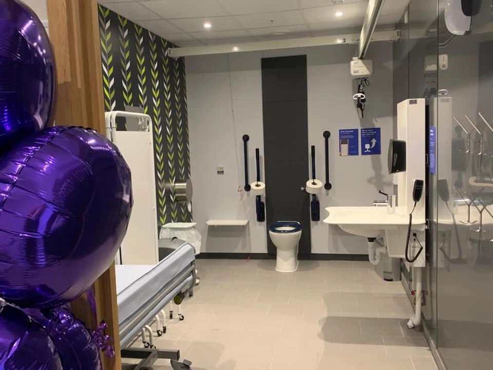 A spacious, accessible public restroom with light grey walls and large purple balloons visible on the left side. The room features a toilet, an adjustable sink, a medical bed/changing table, grab bars, and a ceiling hoist system. The back wall has a decorative black and neon green chevron pattern.