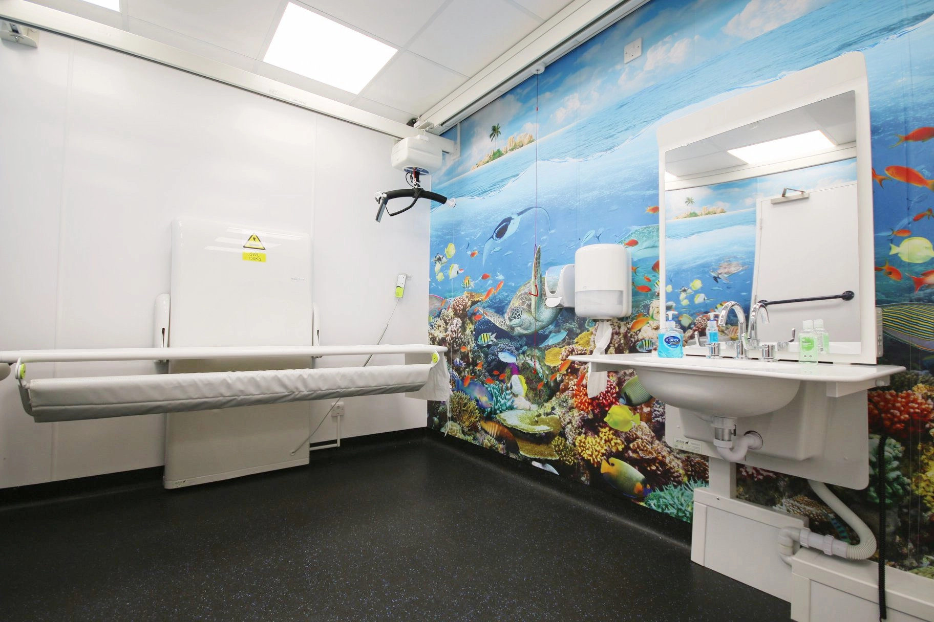 A clean, spacious, accessible public restroom with white walls and dark flooring. One entire wall is covered with a vibrant mural of a colorful underwater ocean scene featuring fish, coral, and a sea turtle. The room includes a height-adjustable changing table, a sink with an extended counter, and an overhead hoist system.