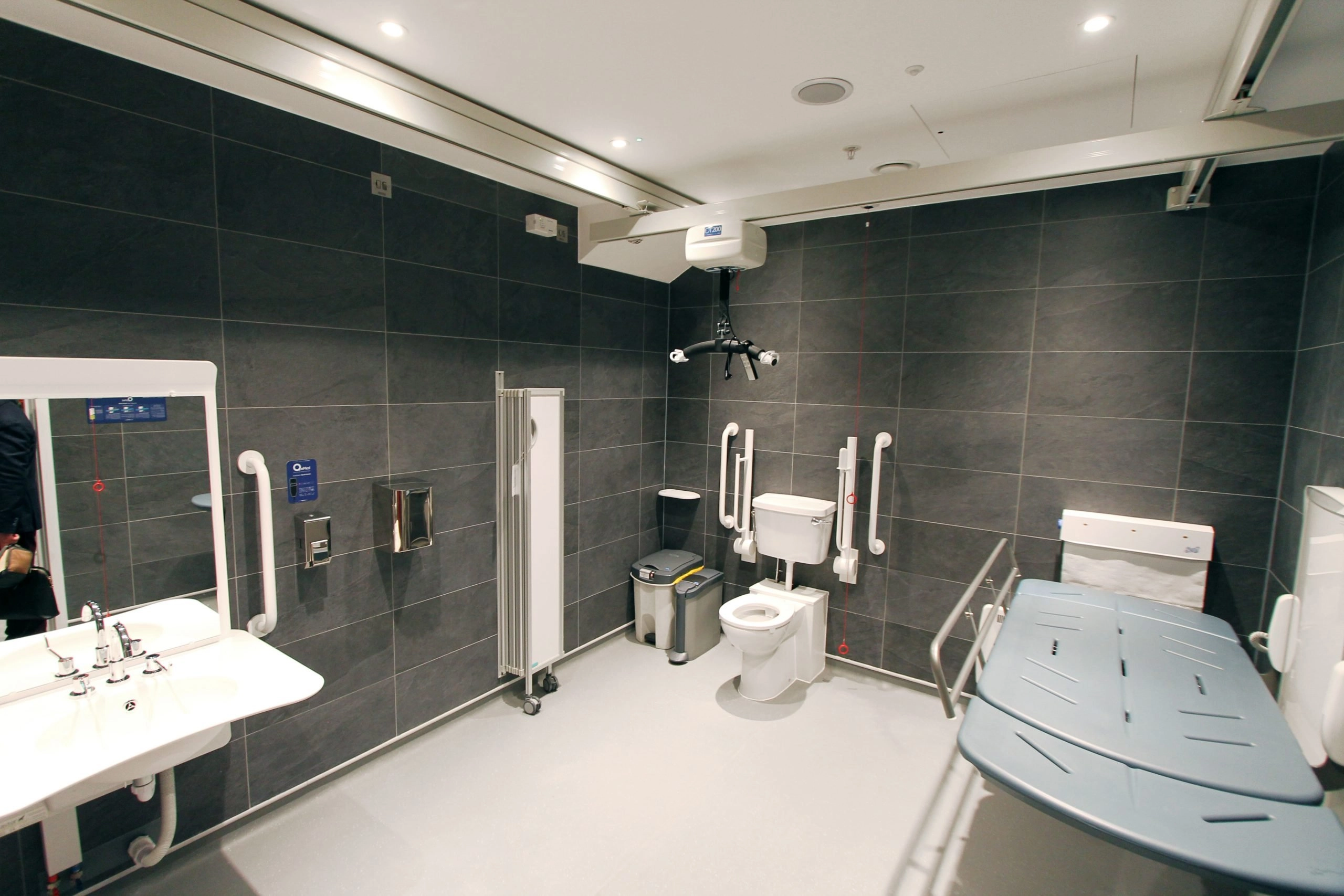 A modern, spacious, fully accessible public restroom (Changing Places style) with large, dark gray tiled walls and a light grey floor. The room includes a toilet with white grab rails, a height-adjustable adult changing table, a wall-mounted sink, and an overhead hoist system.