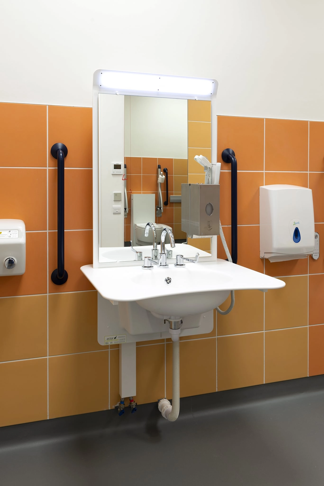 A brightly colored accessible public restroom with orange and yellow square wall tiles. A white sink with an extended basin and a mirror sits in the center, flanked by black vertical grab bars, a paper towel dispenser, and a hand dryer. A toilet is visible in the mirror reflection.