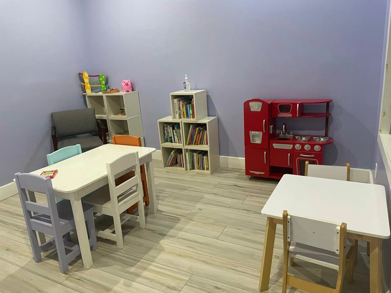 A brightly lit room, likely a play or therapy space for children, with pale lavender walls and light wood-look flooring. The room contains two small white children's tables with colorful chairs, several white cube shelves filled with books and toys, and a bright red, vintage-style wooden toy kitchen set.
