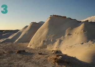 A dramatic landscape of a vast, natural erosion cirque (Makhtesh). The foreground shows the steep, jagged, multi-colored rock walls of the crater, including striking layers of black, red, and yellow earth. In the distance, the wide, flat floor of the crater is visible under a clear blue sky. This is the landscape of the Makhtesh Ramon (Ramon Crater) in the Negev Desert.