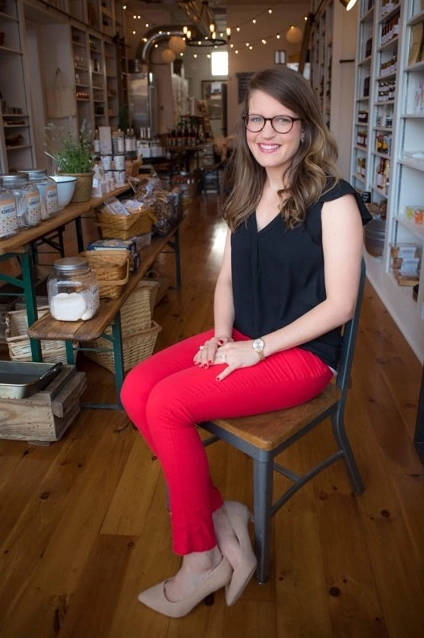 A young woman with glasses and long brown hair, wearing a black top and bright red pants, is seated on a simple wooden chair in the aisle of a well-stocked gourmet or grocery store. She is smiling directly at the camera. The store features wooden floors, industrial lighting, and shelves stocked with various goods.