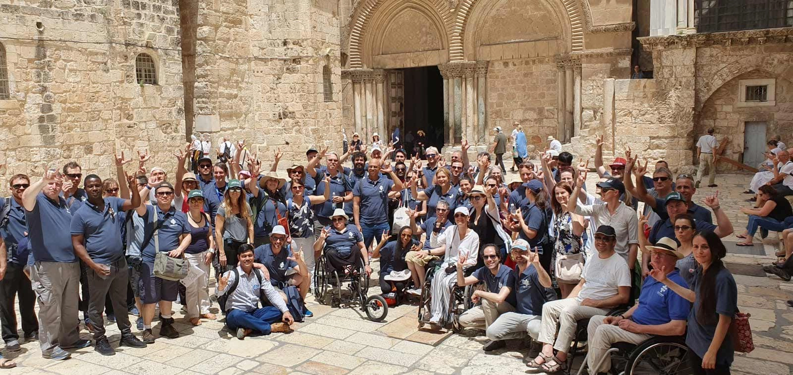 A very large group of people, many wearing matching dark blue shirts and caps, posing together for a photo in a sunny, ancient stone courtyard outside a large church entrance. Several individuals in the group are using wheelchairs. Many people are raising their hands in a celebratory gesture. The stone walls and the dark arched entrance provide a historic backdrop.