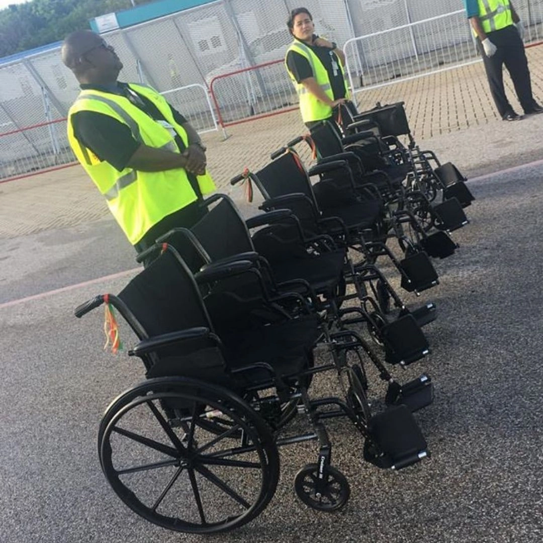 Two uniformed airport or port staff members, wearing safety vests, stand beside a line of several black, manual wheelchairs neatly arranged on an asphalt service area. The male worker in the foreground is looking up, while the female worker stands slightly behind the line of chairs.