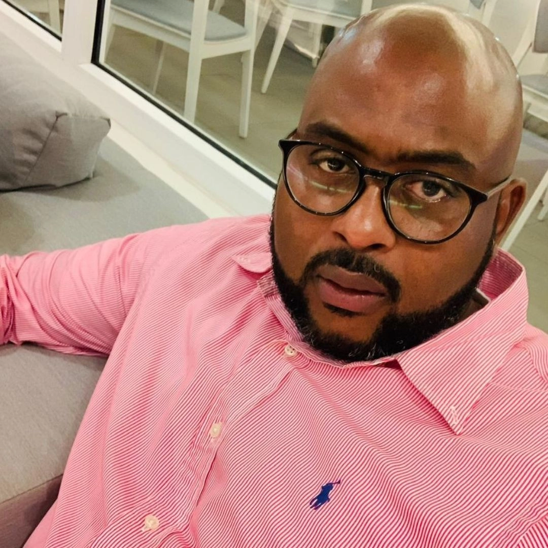 A close-up headshot of a bald, dark-skinned man with a beard, wearing large, dark-rimmed glasses and a pink-and-white striped collared shirt. He is sitting on a grey couch and looking directly at the camera.