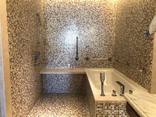 A large, luxurious, accessible bathroom with walls and floor entirely covered in small brown, beige, and white mosaic tiles. The space features a walk-in shower area with a built-in bench, a mounted shower head, grab bars, and a deep soaking bathtub.