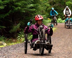 On a dirt path in a forest, a woman in a 3-wheeled low-rise bike grins as cyclists follow behind.