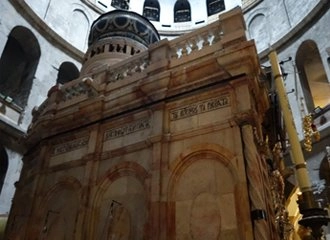 A low-angle interior view of the Aedicule, the structure housing the Tomb of Jesus, inside the Church of the Holy Sepulchre in Jerusalem. The structure is made of pinkish marble with carved inscriptions, and the dark, round dome of the Rotunda is visible above.