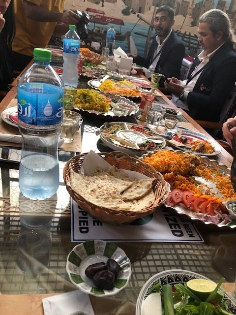 A long table covered with a feast of Middle Eastern dishes, including large platters of saffron-colored rice (Mandi or Kabsa style), a basket of flatbread, a bowl of fresh salad, and a small dish of dates. Two men in dark blazers are seated on the far side, and a large bottle of Arwa water is in the foreground.