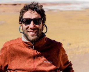 A man with wind-swept hair and sunglasses smiles. Sand stretches out behind him.