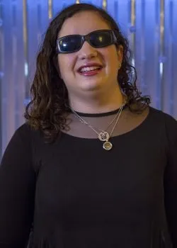 A portrait of a woman with dark, curly hair wearing sunglasses and a black top with a sheer mesh neckline, smiling at the camera. She is wearing two layered silver necklaces with round pendants, and is standing in front of a blurred, shimmering blue and white background.
