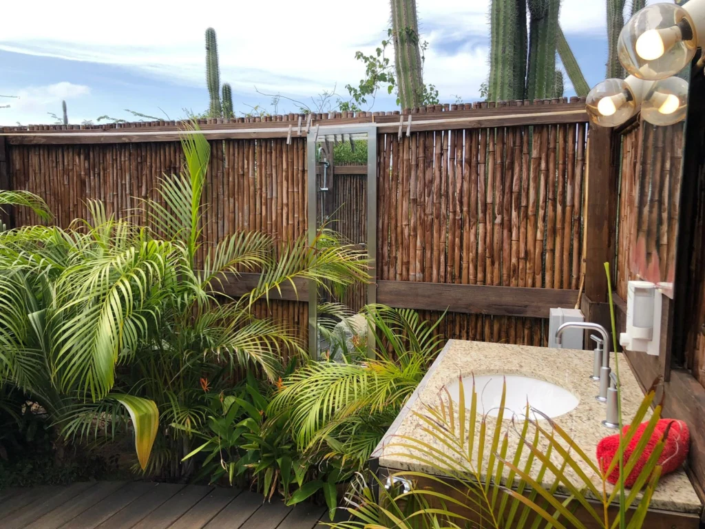 A close-up view of an outdoor bathroom or sink area with a tropical, private feel. The space features a small, light granite-topped vanity with a white basin and a modern faucet. The area is enclosed by a tall bamboo privacy fence, and is surrounded by lush green palm fronds and other tropical plants. Tall cactus plants are visible over the top of the fence, and a modern light fixture with multiple glass globes is on the right.