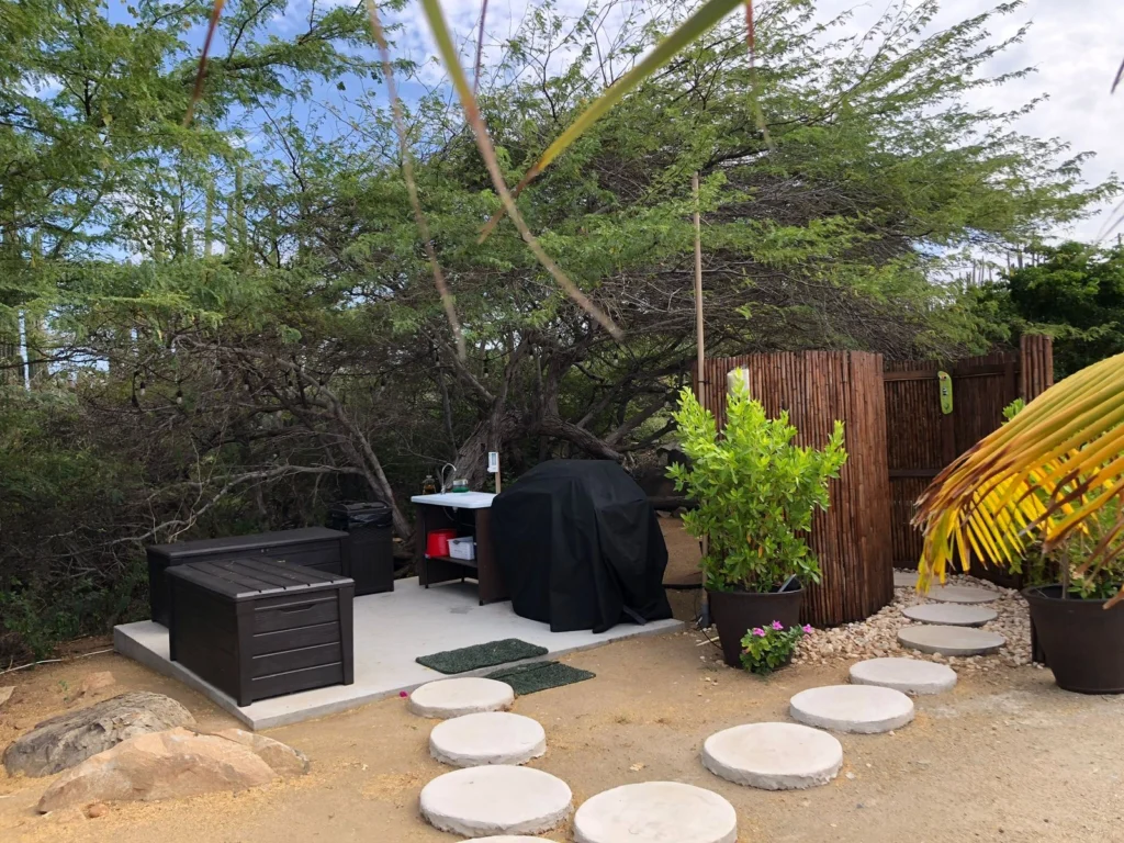 An outdoor cooking and shower area within a glamping setup, nestled under the shade of large, sprawling trees. The area includes a black covered barbecue grill, a small prep table, and black plastic storage boxes set on a concrete pad. A bamboo privacy screen for an outdoor shower is visible on the right, and a path of round concrete stepping stones leads across the dirt ground.