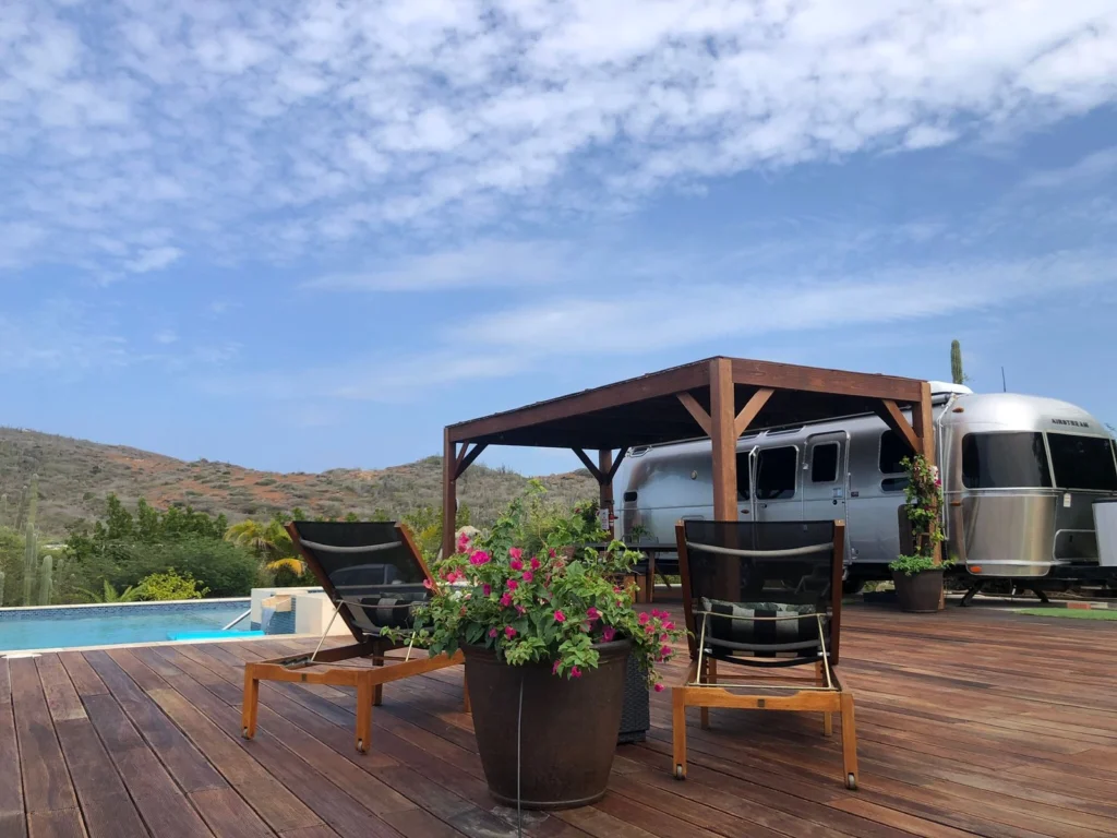 An outdoor scene featuring two wooden and black mesh lounge chairs facing away from the camera, flanking a large potted pink flowering plant. The chairs are positioned on a wide wooden deck that leads back to a small, dark wooden pergola, beneath which a silver Airstream travel trailer is parked. In the background, there is a small swimming pool and a dry, scrub-covered hillside under a bright blue, partially cloudy sky.