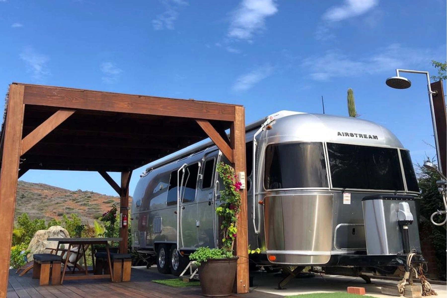 An outdoor scene featuring two wooden and black mesh lounge chairs facing away from the camera, flanking a large potted pink flowering plant. The chairs are positioned on a wide wooden deck that leads back to a small, dark wooden pergola, beneath which a silver Airstream travel trailer is parked. In the background, there is a small swimming pool and a dry, scrub-covered hillside under a bright blue, partially cloudy sky.