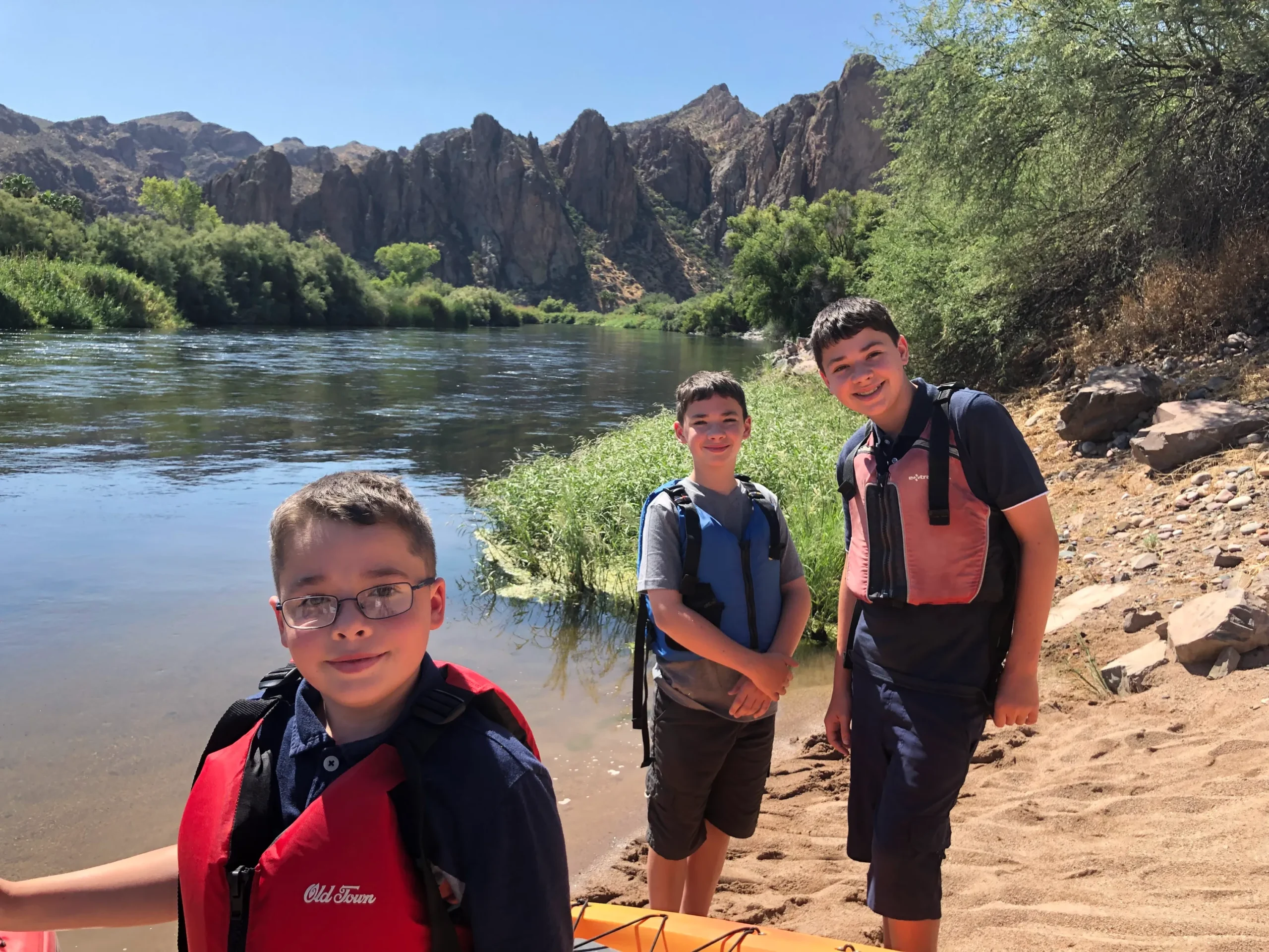 Three boys stand smiling on a sandy riverbank on a sunny day. They are all wearing life jackets—the one in the foreground has a red life jacket, and the other two have blue/black and dark red/black life jackets. Behind them is a calm river lined with tall green reeds and trees. In the distance, large, rugged, dark mountains and rock formations rise sharply under a clear blue sky.