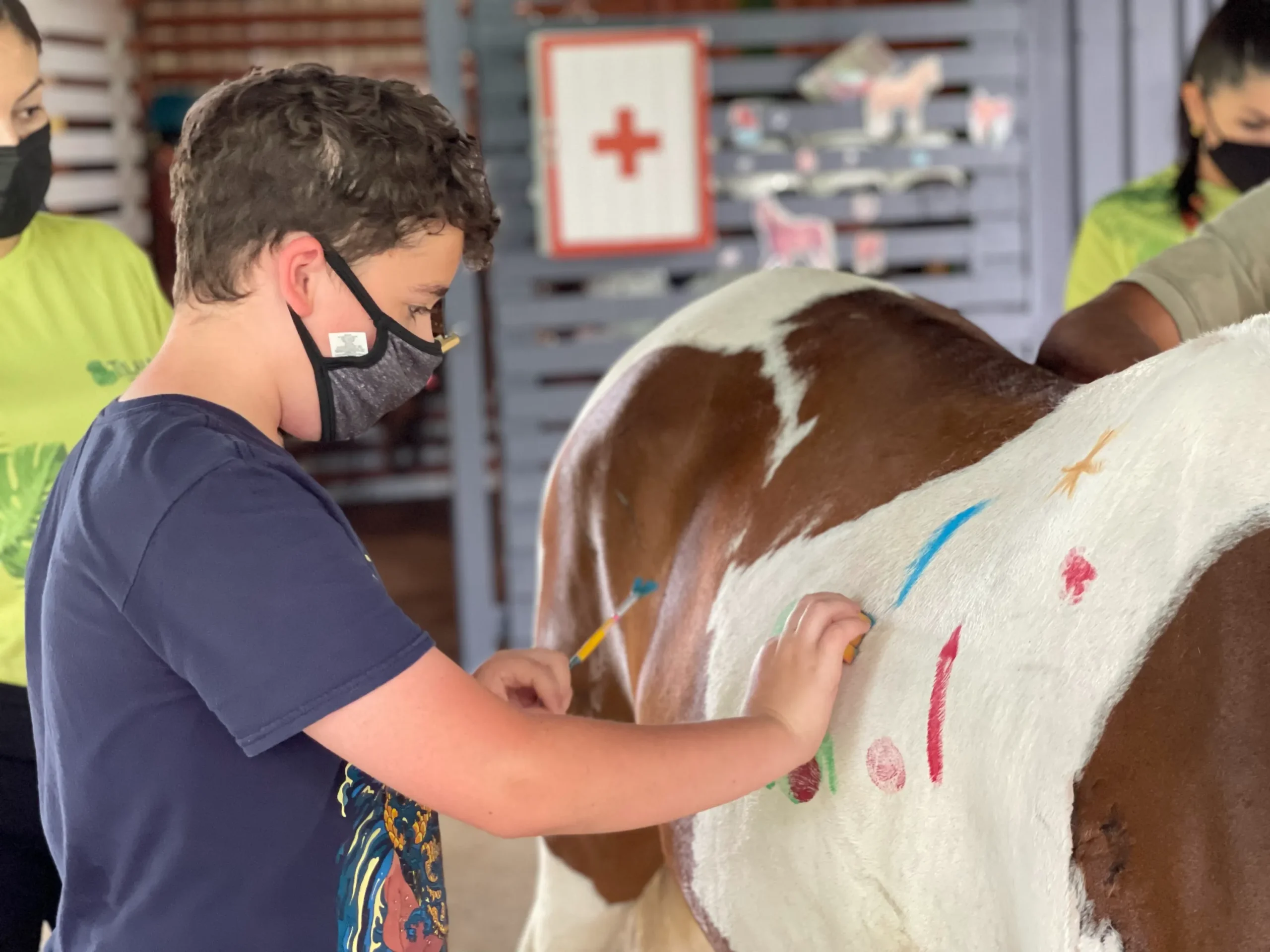 A young boy wearing a dark t-shirt and a protective face mask is focused on painting a design onto the white flank of a horse. He is holding a small brush and applying colorful, non-toxic paint. The horse is a brown and white pinto, and the activity appears to be part of a therapy or educational program, possibly involving creative expression. In the blurred background, other people wearing masks and a sign with a red cross medical symbol are visible.