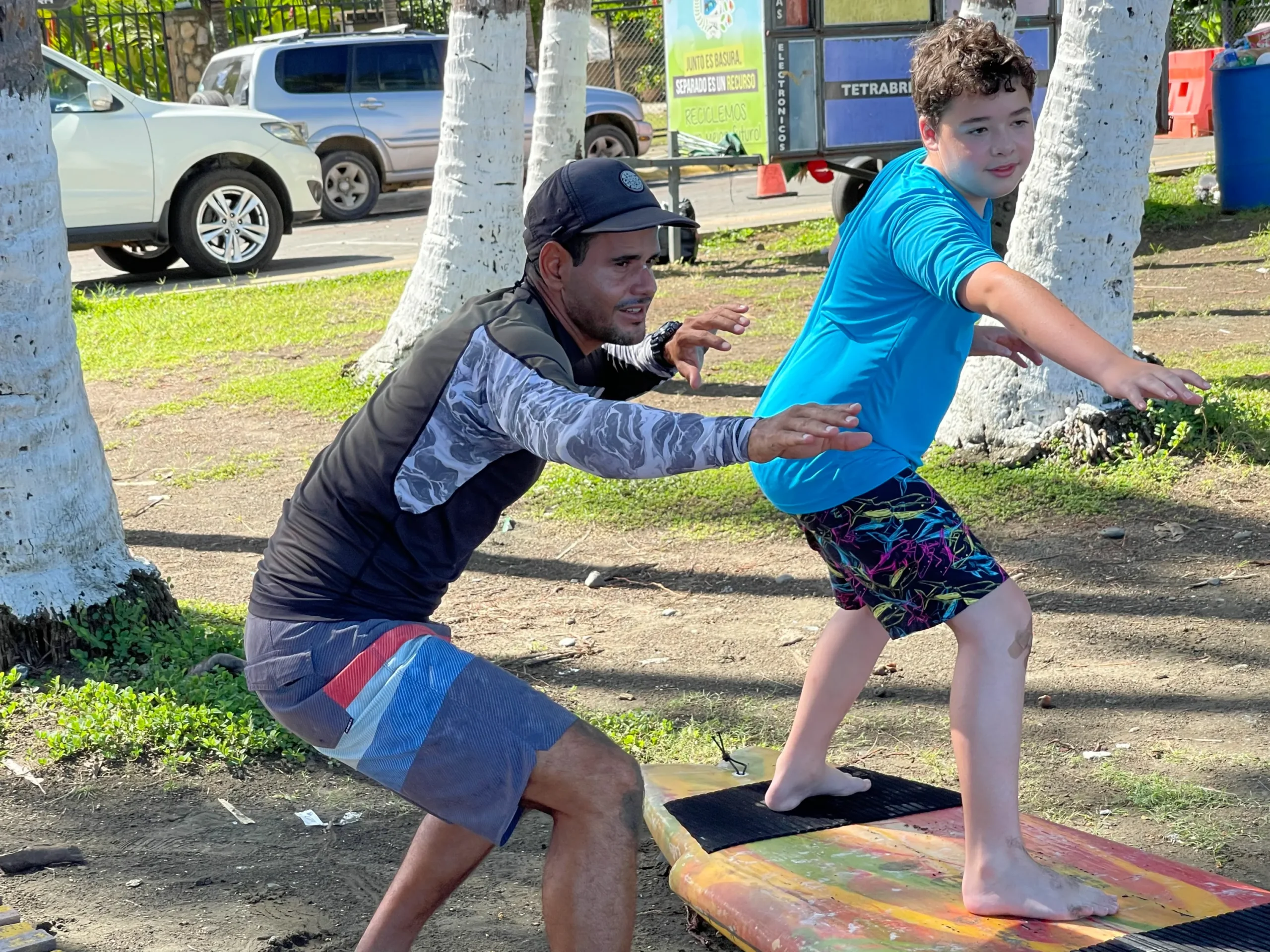 An instructor, a man wearing a dark rash guard and board shorts, is demonstrating a surfing or balance technique on land to a young boy. Both are standing on a large, colorful surfboard or balance board placed on the grass. The instructor is in a low, wide stance with his arms out, coaching the boy who is trying to mimic the posture. They are outdoors, with white-trunked trees nearby and several parked cars and a trailer visible in the background on a sunny day.