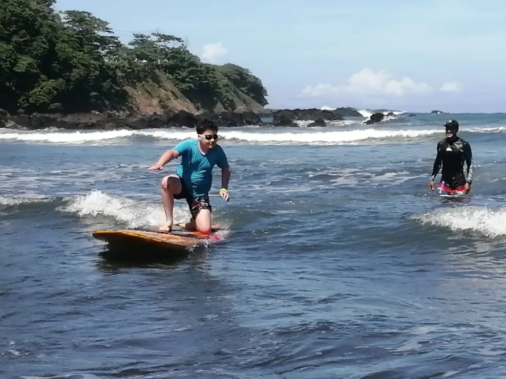 Under a clear sky, a boy surfs in the ocean, a rocky crag with breaking waves in the distance.