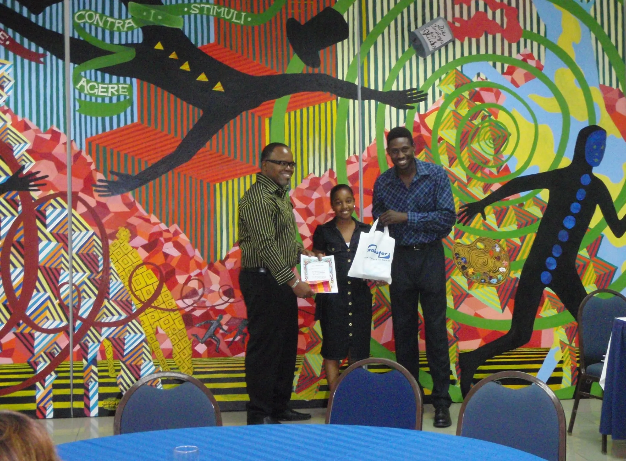 Three people—an older man, a young woman, and a young man—are standing and smiling in front of a colorful, abstract mural wall. The older man, on the left, is wearing a striped green and black shirt and is holding a certificate. The young woman in the center is holding a white shopping bag and a certificate. The young man on the right is wearing a checkered shirt. A blue tablecloth is visible in the foreground with dark chairs around it.