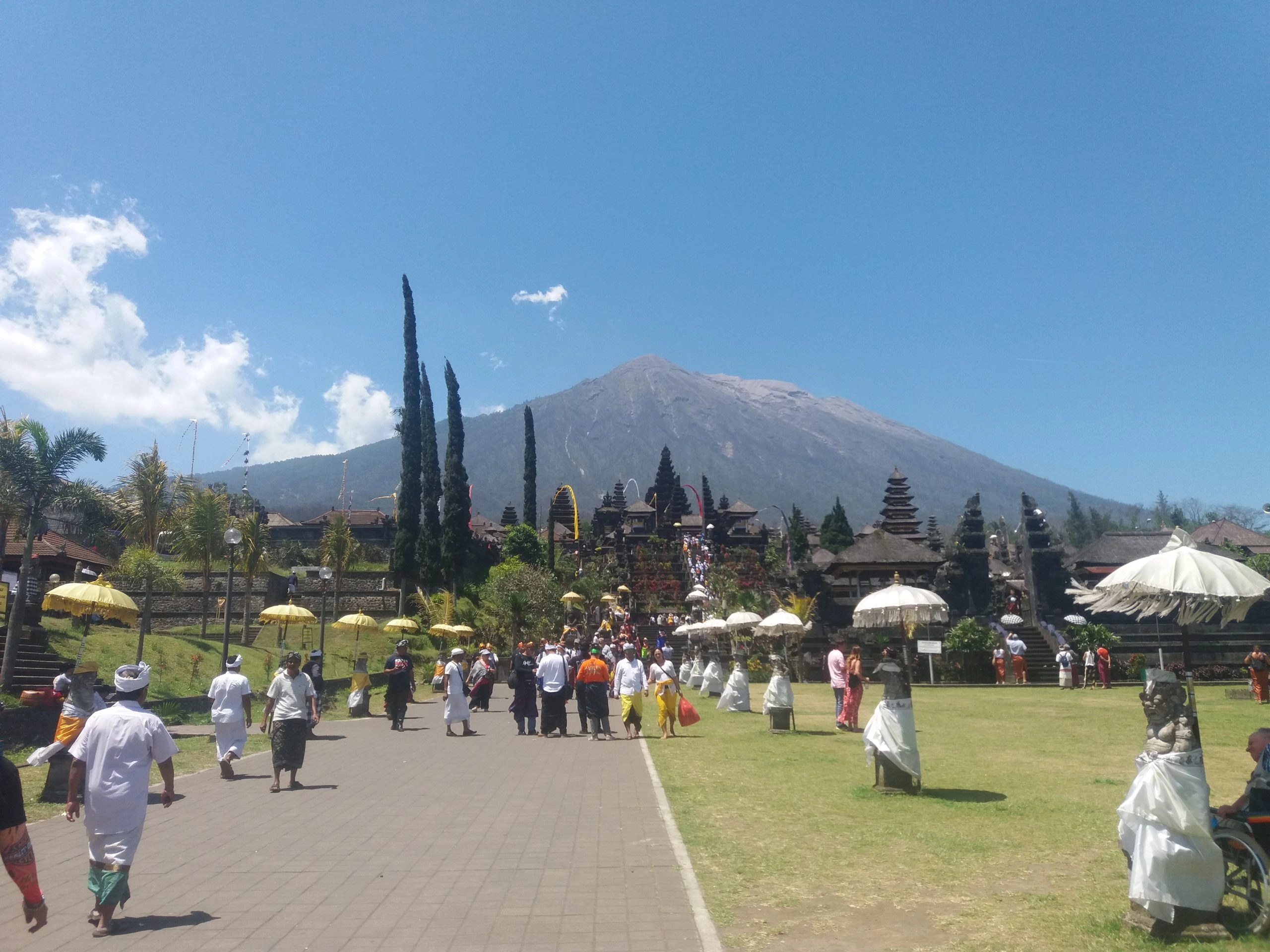A wide, sunny shot of the Pura Besakih temple complex in Bali, Indonesia, with the majestic Mount Agung volcano rising in the background under a clear blue sky. A wide paved courtyard leads toward the temple, where many people in traditional Balinese white and yellow attire are walking.