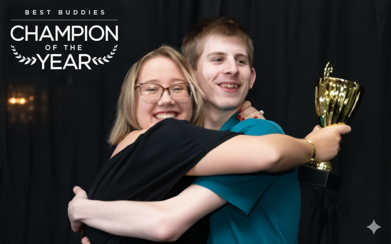 A young woman with glasses and a young man are embracing and smiling at the camera. The man is holding a large golden trophy. The image has a graphic overlay in the upper left corner that reads "BEST BUDDIES CHAMPION OF THE YEAR." The background is dark.