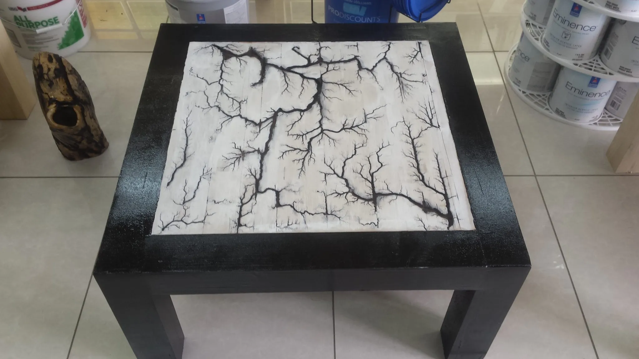 A square wooden coffee table with a black painted frame. The white tabletop insert features a striking pattern of black, branching, tree-like burn marks created using the Lichtenberg figure technique. The table is resting on a tiled floor in a store setting.