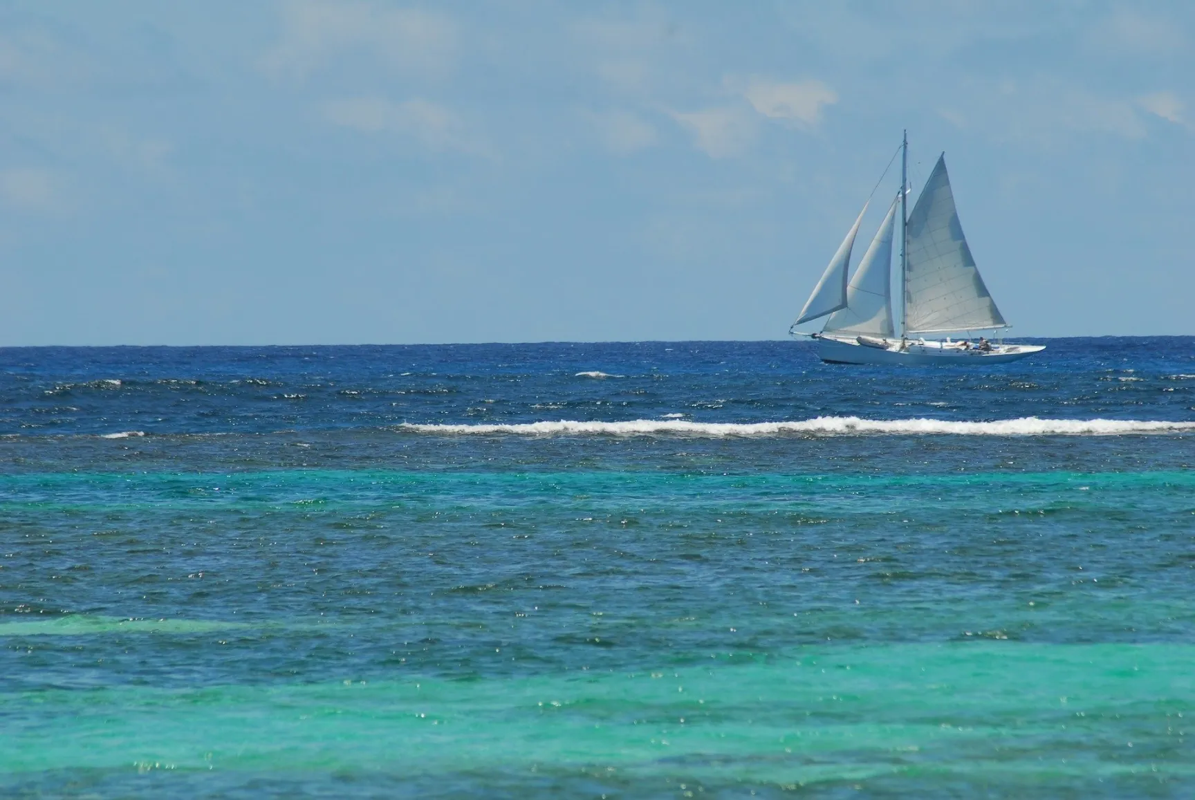 A wide-angle view of the ocean with distinct bands of color, showing a turquoise and aqua blue foreground over a reef, transitioning to a dark, deep blue open sea. A white sailboat with two sails is visible sailing on the horizon beneath a light blue sky with scattered clouds.