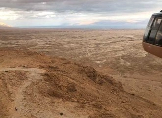 A view from a cable car looking down over the vast, rugged, brown desert plateau below Masada. The Dead Sea and distant mountains are visible on the horizon under a cloudy sky. The right side of the image shows the bottom edge of the cable car cabin.