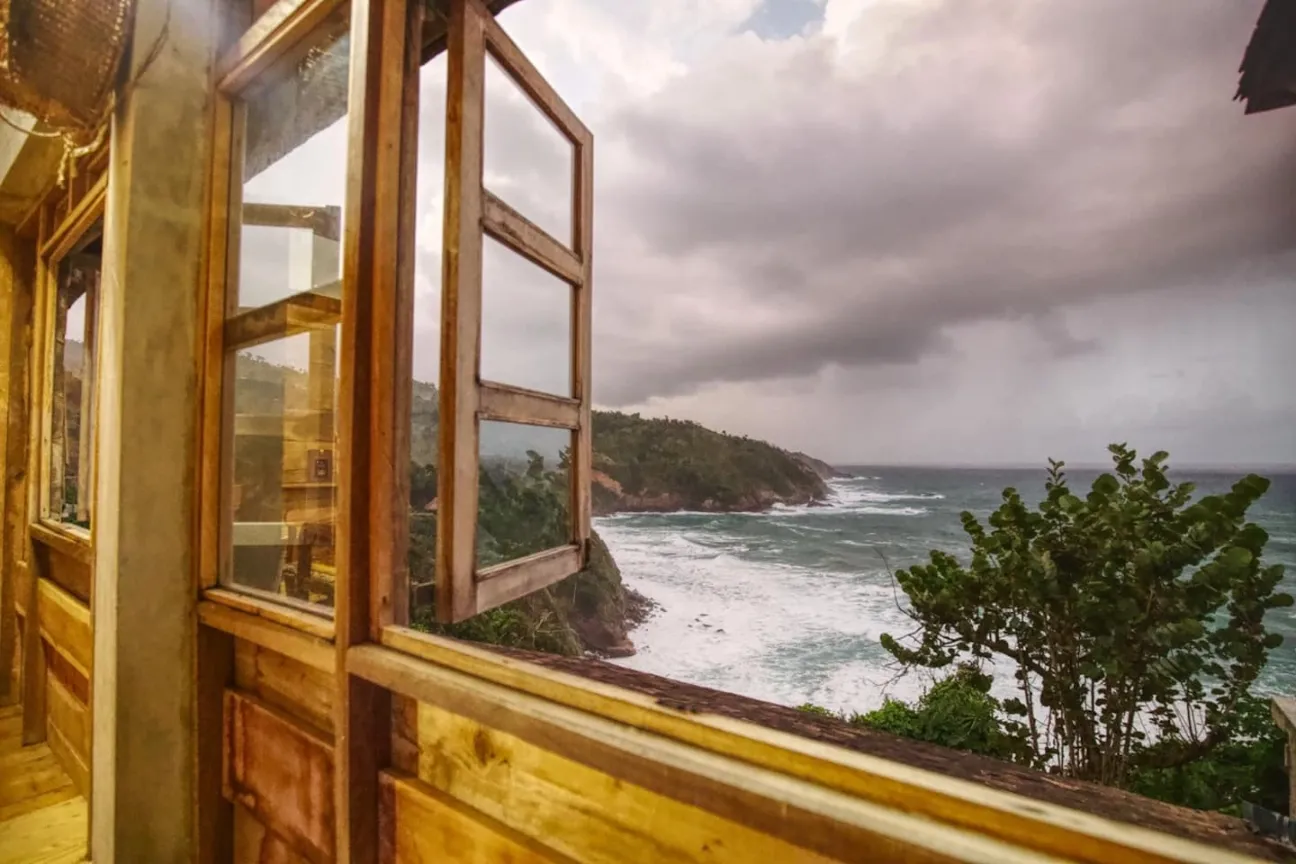 A view of a dramatic, rough coastline and deep blue ocean seen from an open wooden-framed window inside a wooden cabin. Large, white waves crash against the rocky green cliffside under a dark, stormy, overcast sky.