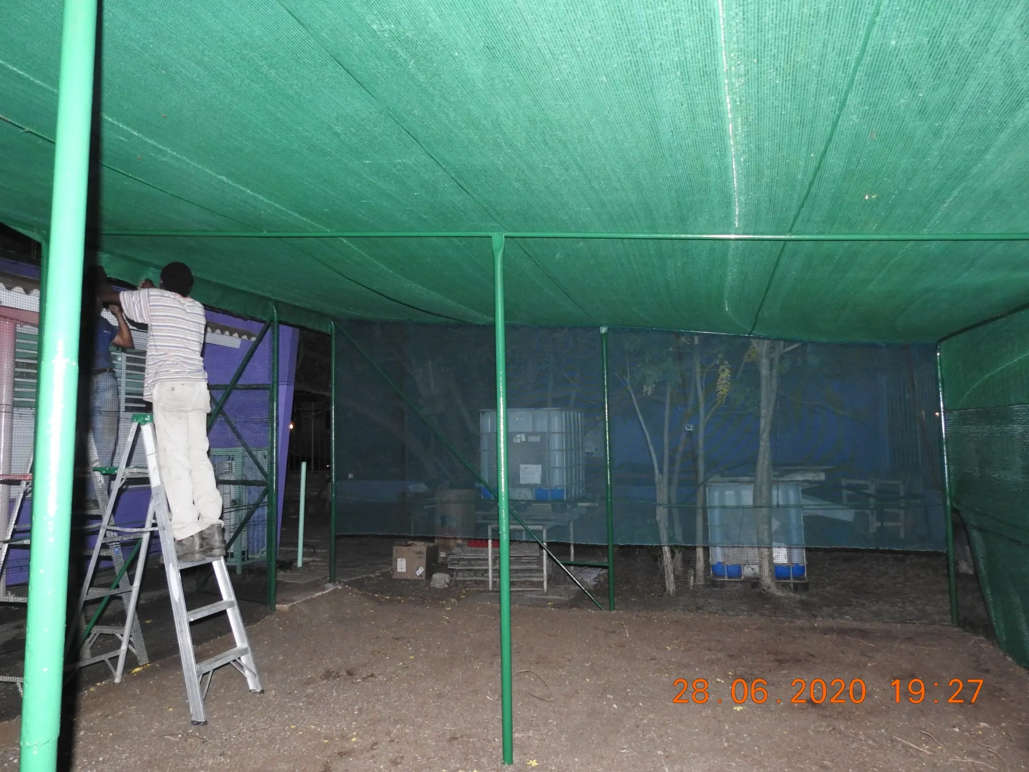 The interior of a newly constructed shelter or grow house framed with green metal poles and covered with a green shade cloth or netting. A person wearing a striped shirt and tan trousers is standing on a step ladder, working on the ceiling near the left side. Large white plastic water tanks are visible through the back netting. The date stamp "28.06.2020 19:27" is in the bottom right corner.