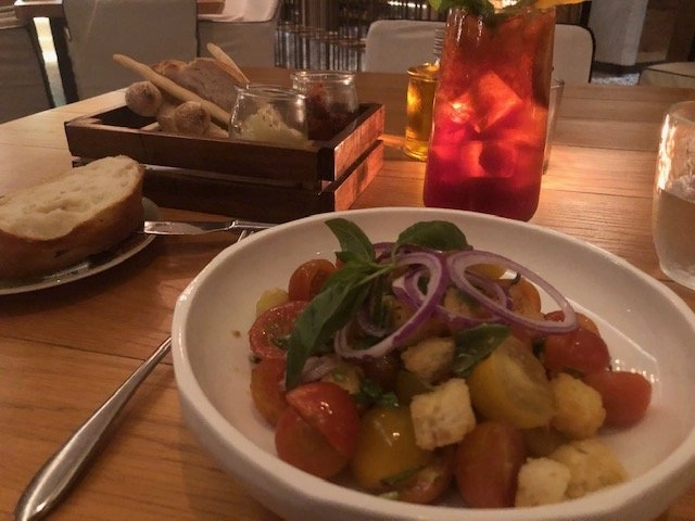 dimly lit restaurant table setting featuring a white bowl containing Panzanella salad, which is made of chopped cherry tomatoes, bread croutons, red onion, and fresh basil. In the background, there is a dark wooden box with various types of bread and breadsticks, and a tall glass with an iced red cocktail.