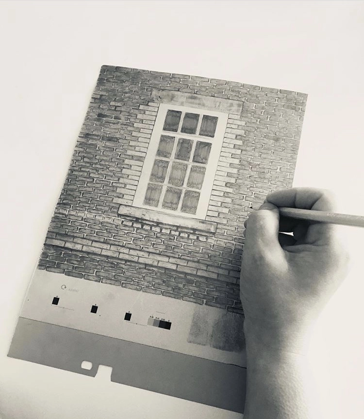 A black and white photograph of a hand sketching a detailed window set within a brick wall onto a piece of paper. The paper also features a large, empty gray area at the bottom with small, dark square markings.