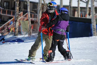 An adaptive skiing lesson is taking place on a sunny, snow-covered slope. A person in a red jacket, likely an instructor, stands next to a skier wearing a purple jacket and black pants. The skier is using adaptive equipment, including specialized outriggers attached to their hands for balance and support. Both individuals are wearing helmets and goggles. Buildings and trees are visible in the blurred background.