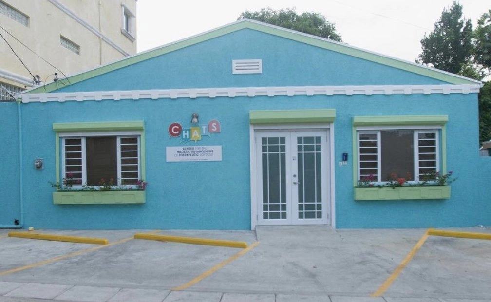 The exterior of a one-story building painted a bright turquoise blue with light green trim around the windows. The front has a central white double door and two windows with white grilles and green window boxes containing pink flowers. Above the door is a sign with the logo "CHASS" and the text "CENTER FOR HOLISTIC ALLYSHIP & THERAPEUTIC SERVICES." The foreground shows a paved parking area with yellow-painted parking stripes.