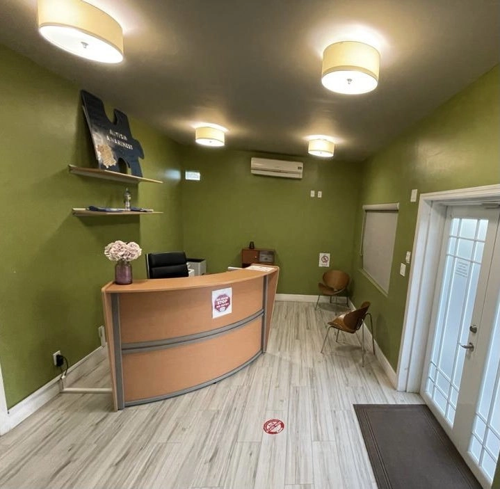 The interior view of a reception or waiting area painted in a muted olive green color. A light wood and tan curved reception desk is in the center, with a computer monitor and a vase of pink flowers. Above the desk, two wall shelves hold various items, and a large blue cutout shape with text about Autism Awareness leans against the wall. The floor is light-colored wood-look tile, and several round ceiling lights illuminate the space. Two simple chairs sit near the windows.