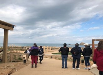 A group of tourists walking on a paved pathway towards the Mediterranean Sea at an archaeological site, likely Caesarea National Park. The walkway is lined with wooden fences and leads to the coastline and water under a partly cloudy sky.