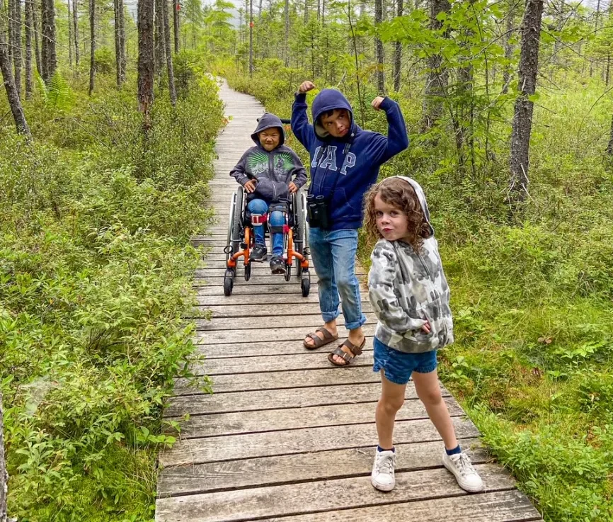 Three kids pose on a wooden walkway in a forest; two standing and one in a wheelchair.