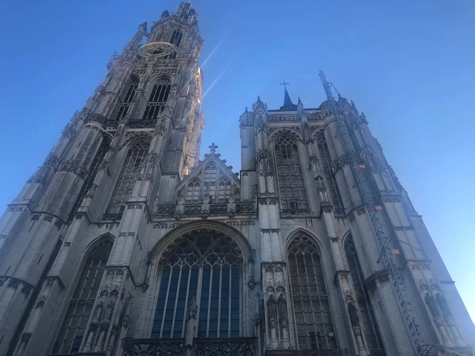 A dramatic low-angle view looking up at the majestic, gray stone facade and tower of a towering Gothic cathedral against a clear, bright blue sky. The main entrance features large stained glass windows, intricate stonework, and multiple levels of arches and spires, characteristic of European Gothic architecture.