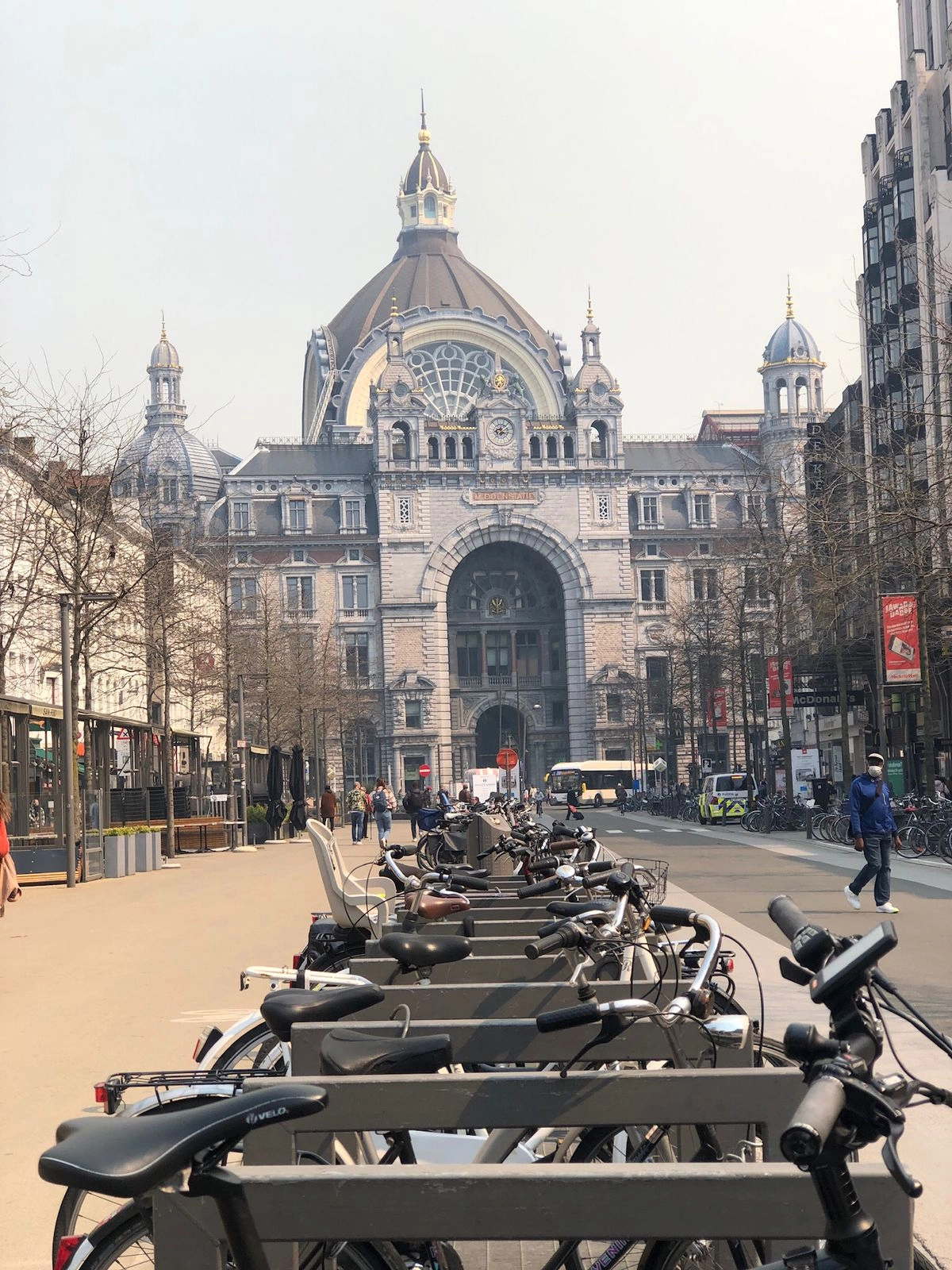 A street view looking toward the grand, ornamental facade of Antwerp Central Station (Antwerpen-Centraal) in Belgium. The station's Neo-Baroque exterior features a large central dome and arched entrance. In the immediate foreground, there is a large, crowded rack of parked bicycles, with an open pedestrian plaza leading toward the building.