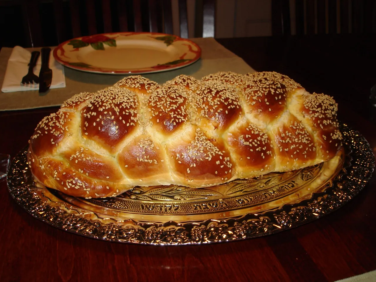 A large, beautifully braided challah bread with a shiny, golden-brown crust, generously topped with sesame seeds. The loaf is presented on an ornate silver-colored platter featuring etched designs and Hebrew text. In the blurred background, a dining table is set with a white and red decorative plate and silverware.
