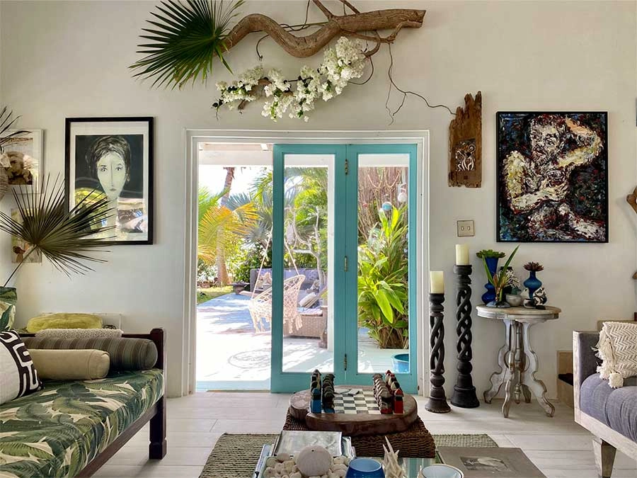 A bright, tropically-themed living room with white walls and floors. The room is decorated with large palm fronds and driftwood, with framed artwork on either side of the turquoise blue French doors that lead out to a sunny patio surrounded by lush green foliage. A daybed with a palm-print cushion and a chess set on a coffee table are visible.